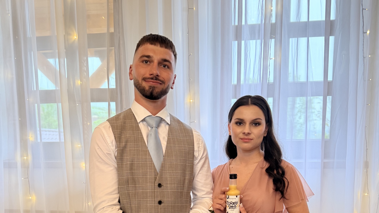 Muž a žena držia fľašu Zdravý Shot v svadobne vyzdobenej sále.